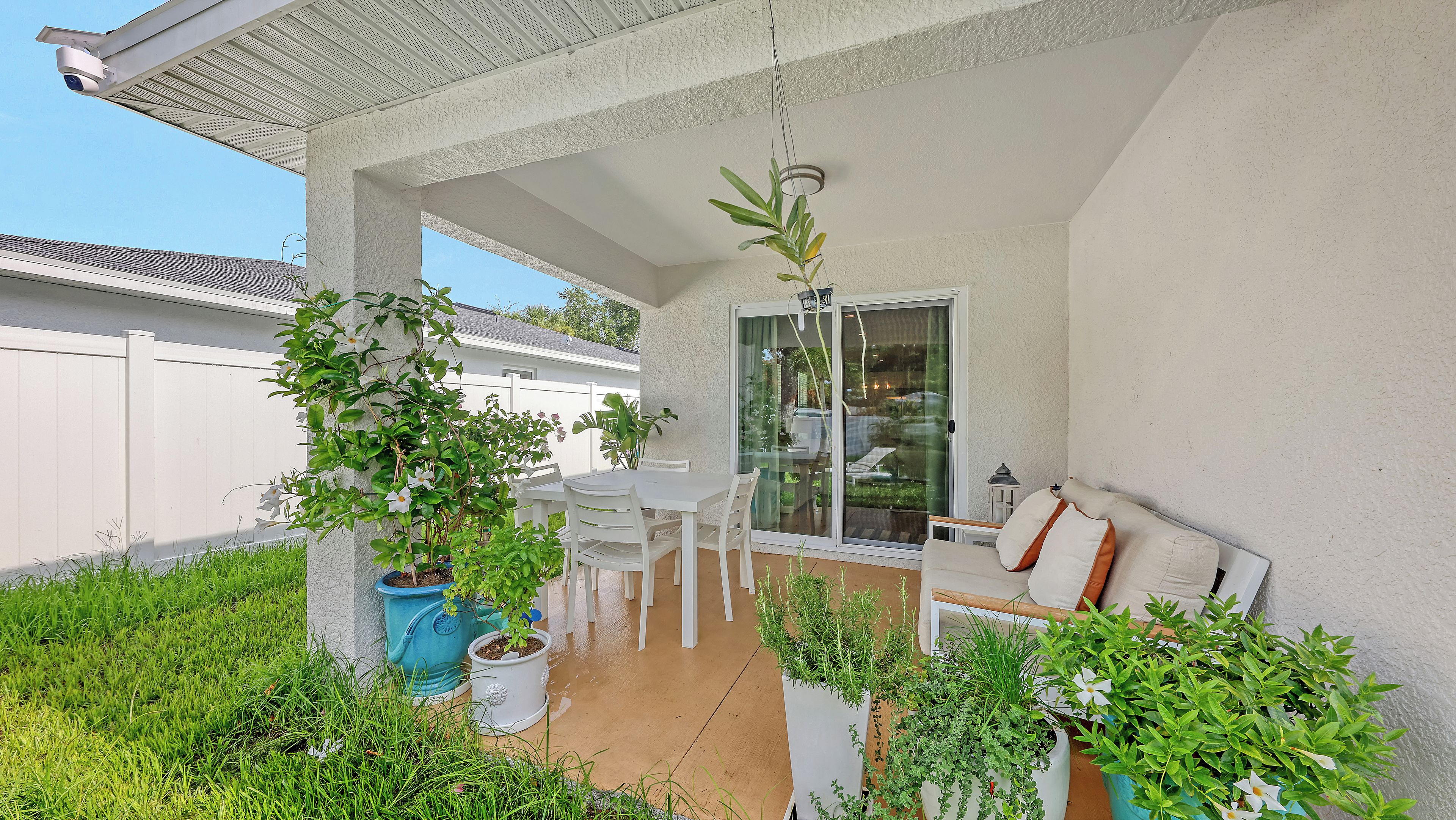 An outdoor patio area with a white table and chairs, surrounded by potted plants and a sliding glass door leading to a glass-enclosed space.