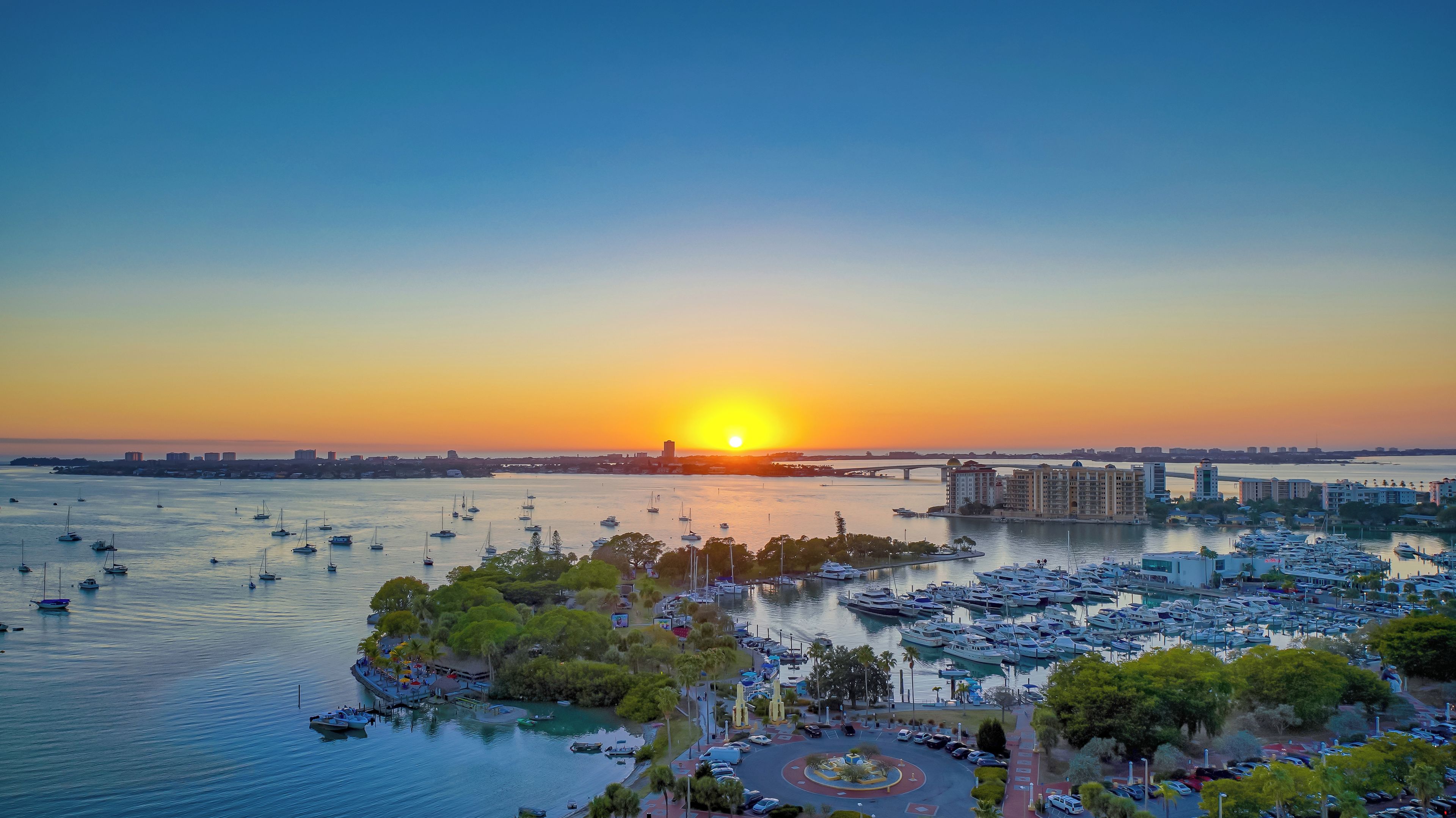 A marina with numerous boats, a bridge, and buildings in the background during sunset.