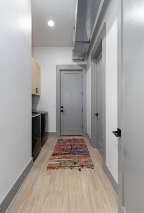 A narrow hallway with a grey door at the end, a colorful rug on the floor, and a small cabinet to the left.