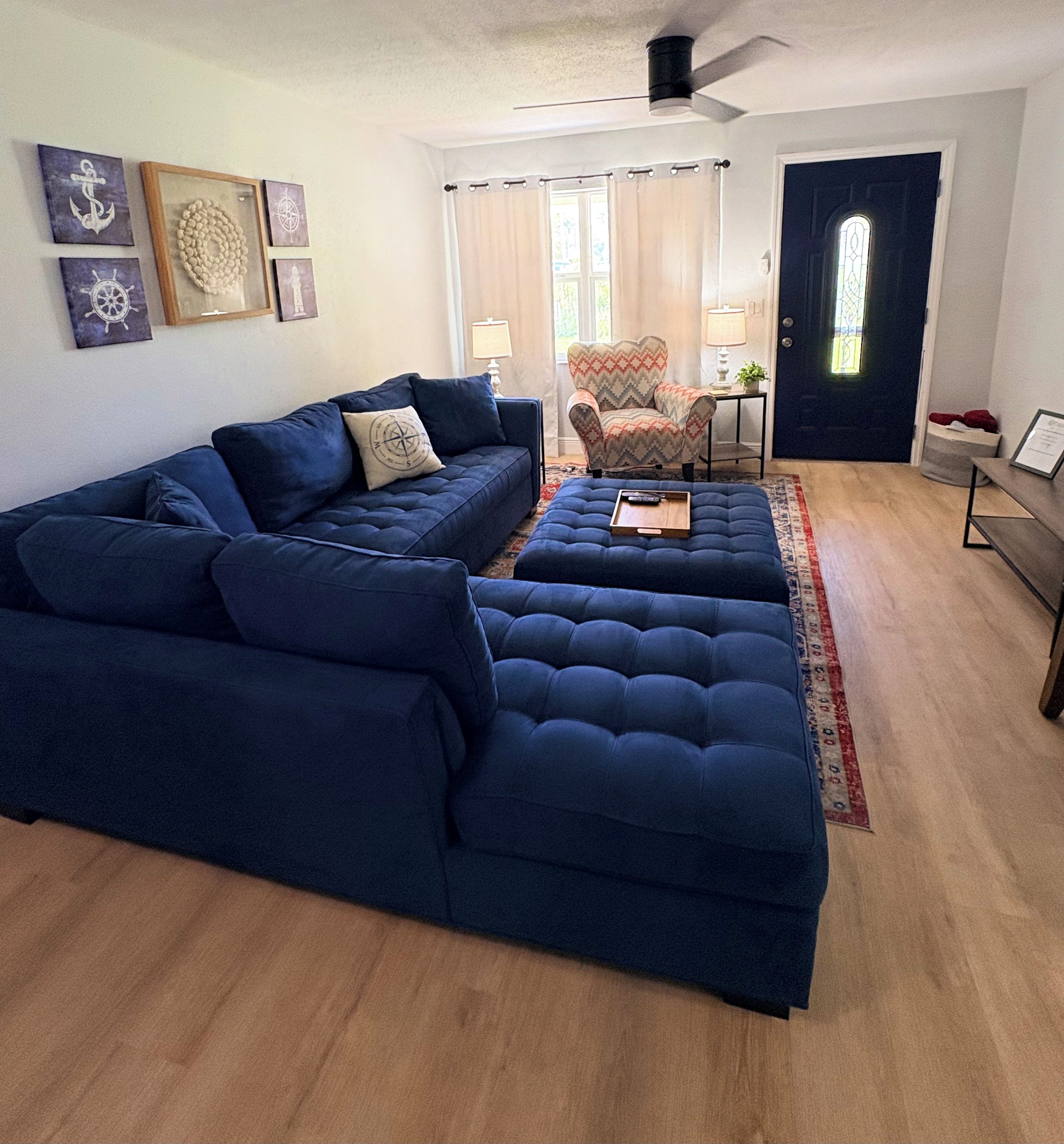 Un salon comprenant un canapé d’angle, un fauteuil à motifs, une petite table avec une lampe et une porte avec une fenêtre.