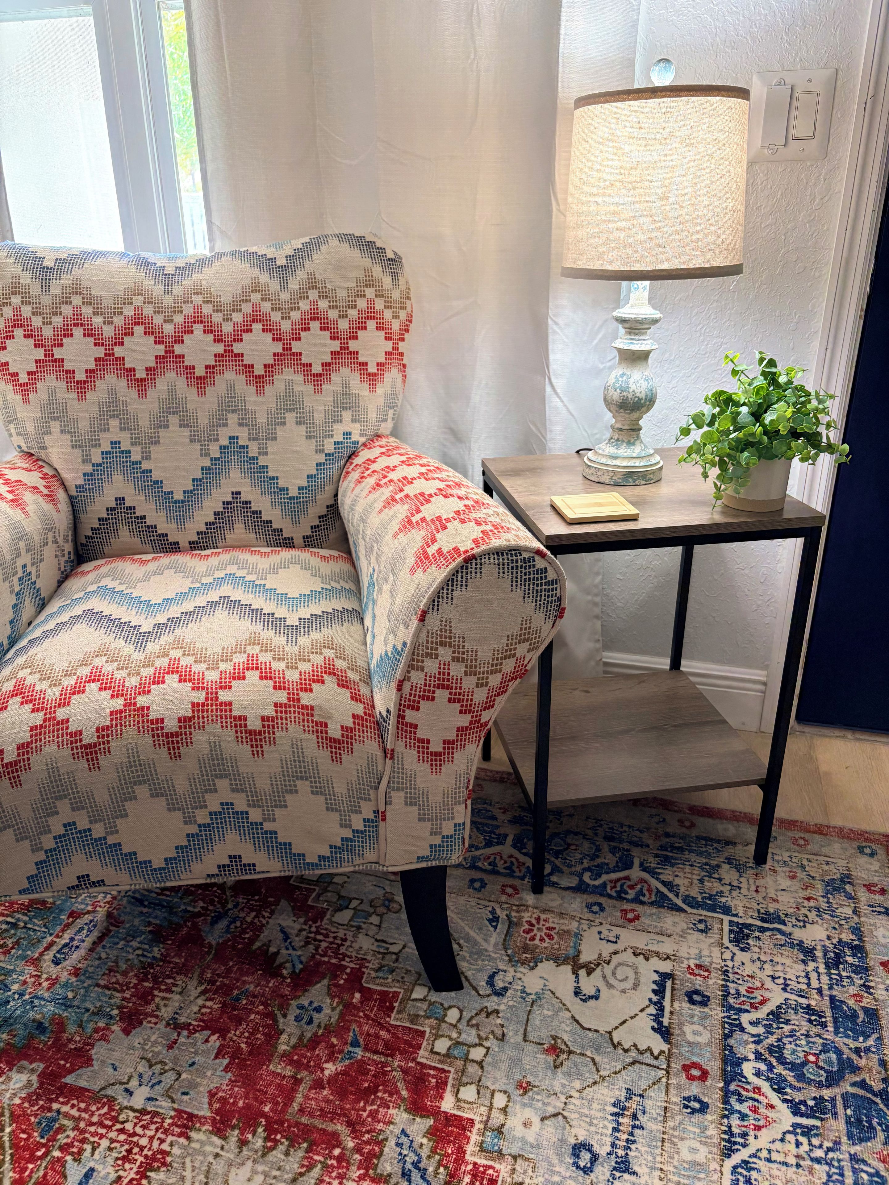 Une pièce avec un fauteuil à motifs, une petite table avec une lampe et une plante en pot.