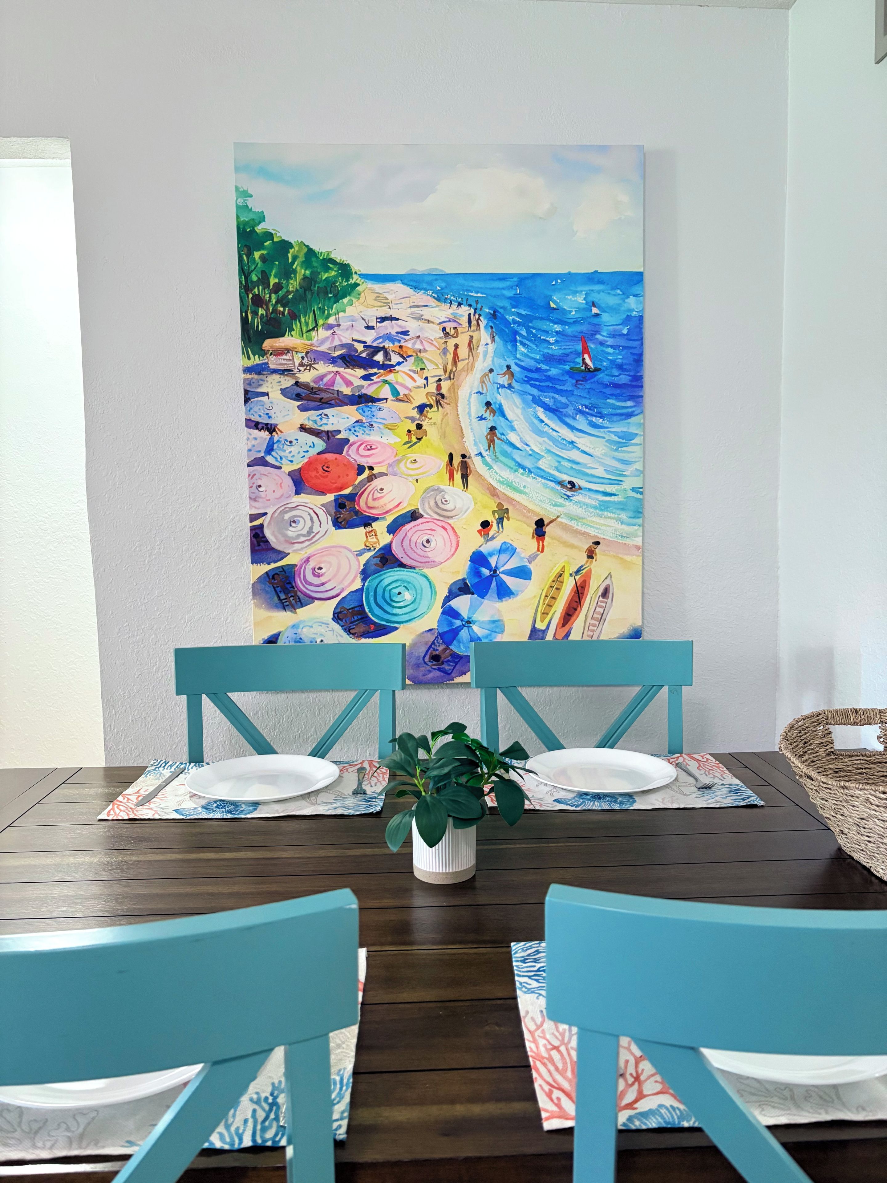 A dining table with chairs, a potted plant, and a colorful beach painting on the wall.