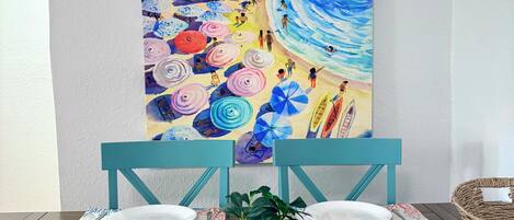 A dining table with chairs, a potted plant, and a colorful beach painting on the wall.