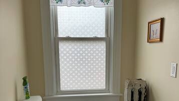 Un baño con inodoro, una ventana con cortina decorativa, un radiador y un cuadro enmarcado en la pared.