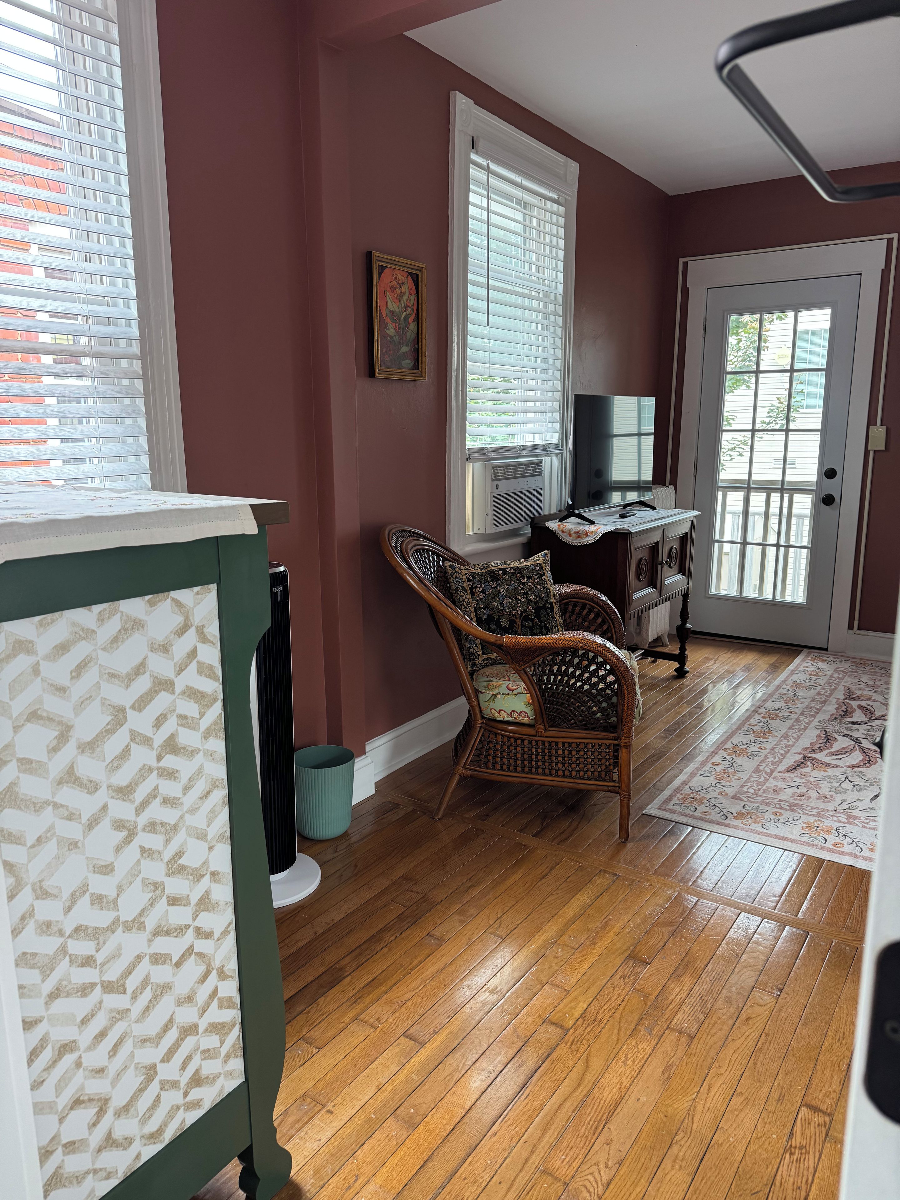 Una habitación con piso de madera, una silla de mimbre, un televisor y una puerta con ventanal.