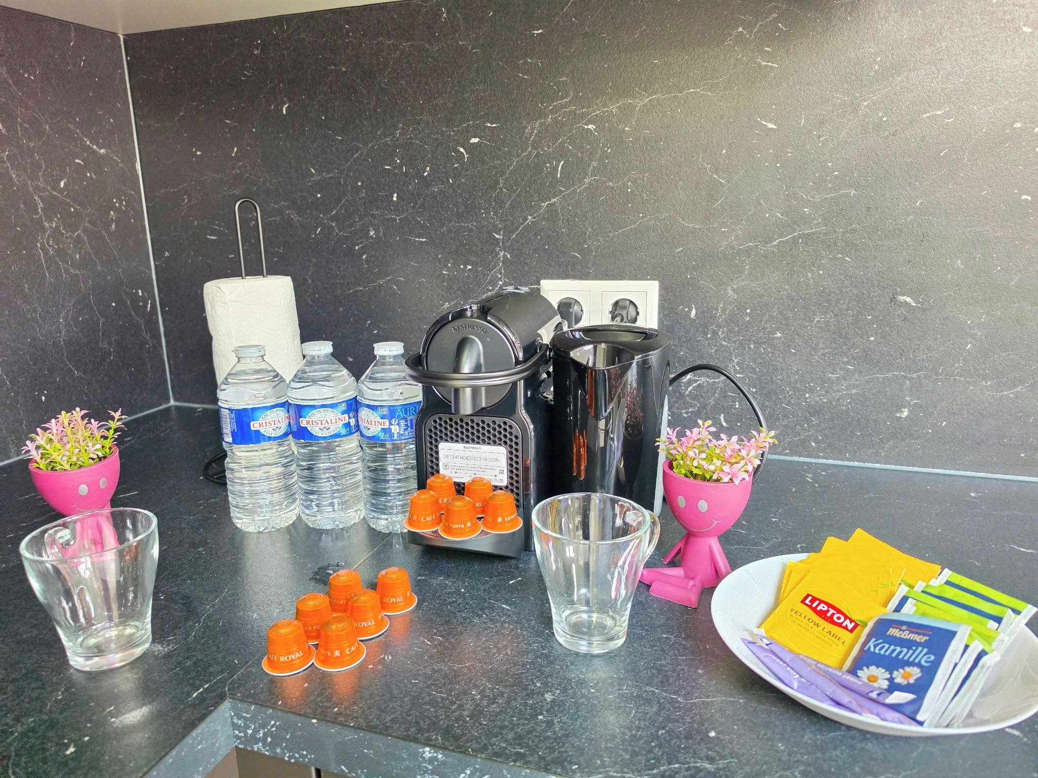 Un comptoir de cuisine avec une machine à café, des bouteilles d’eau, des verres et une assiette contenant des aliments.