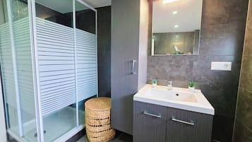 Une salle de bain moderne avec une douche à l’italienne en verre, un lavabo blanc avec un miroir au-dessus et des meubles aux teintes foncées.