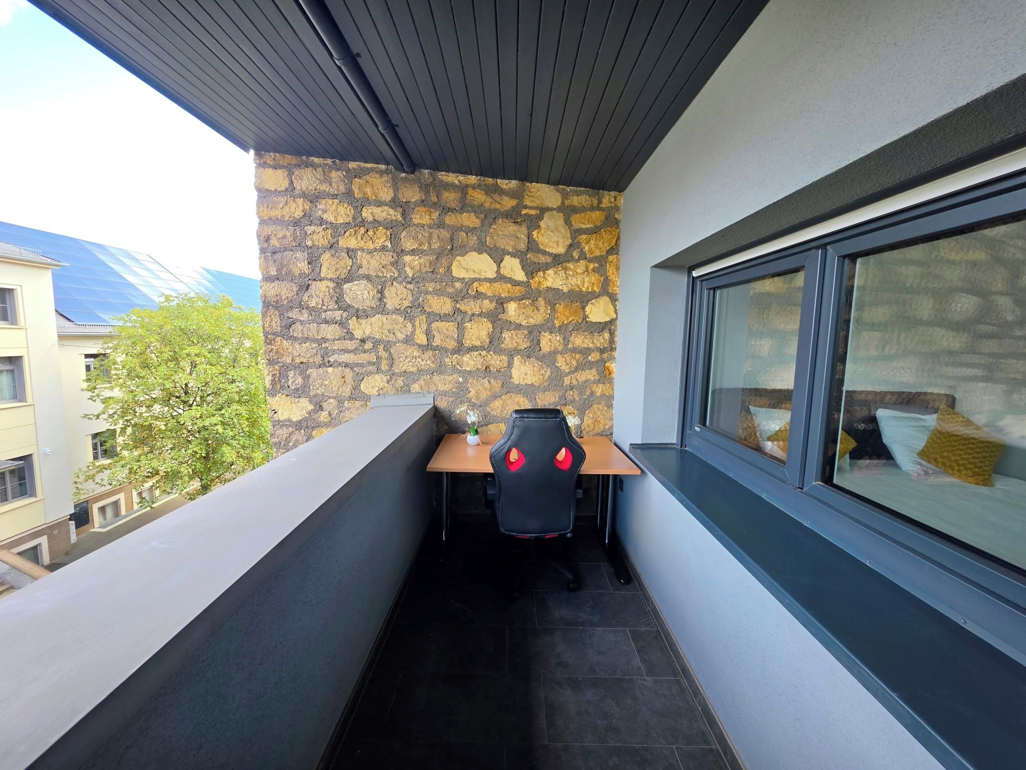 A balcony with a stone wall, a wooden table, and a black chair with red cushions.