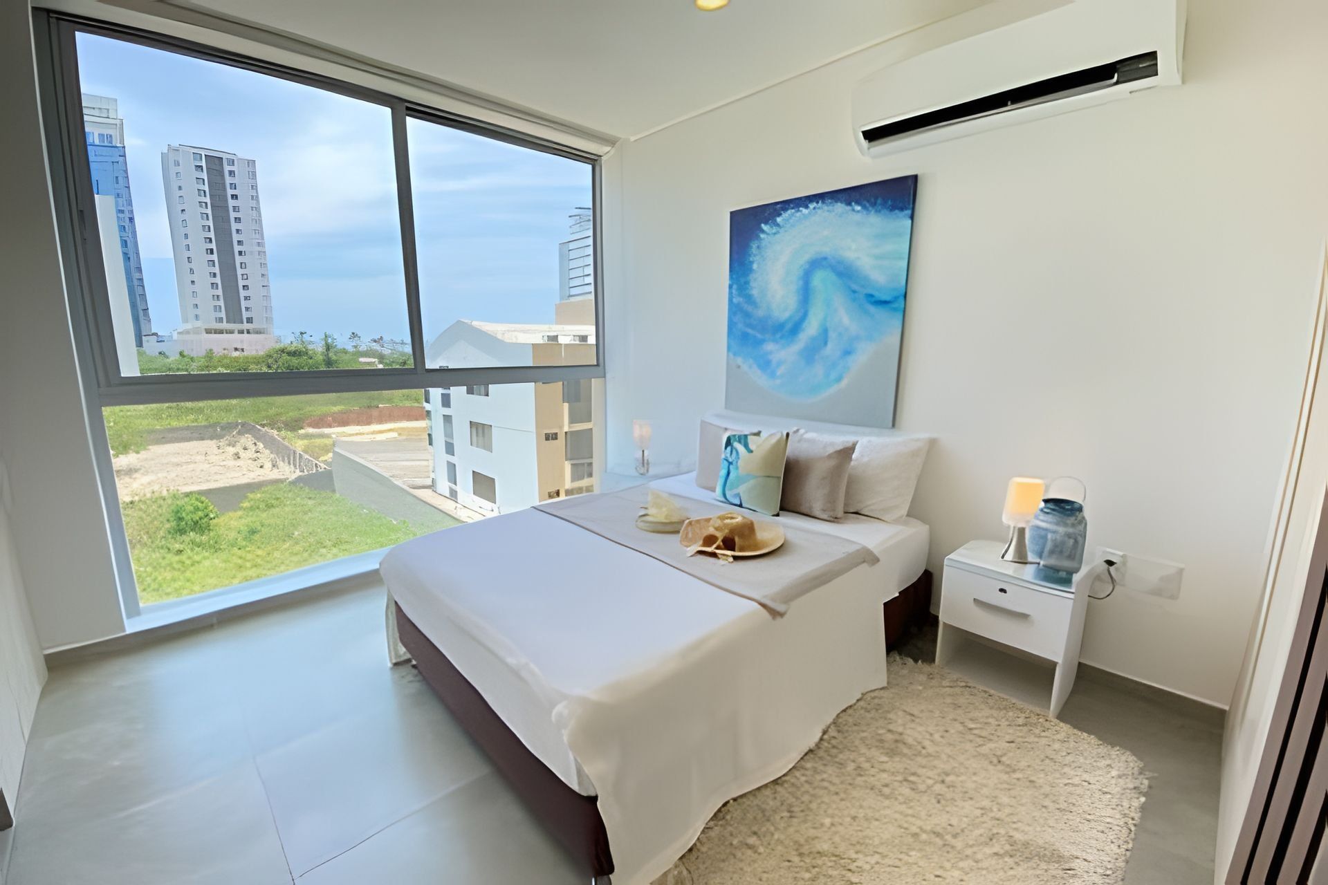 A bedroom with a large window, a bed with white linens, a nightstand with a lamp, and a painting on the wall.