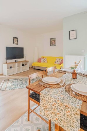 Ein modernes Wohnzimmer mit einem gelben Sofa, einem Esstisch für vier Personen und einem Flachbildfernseher auf einem Standfuß.