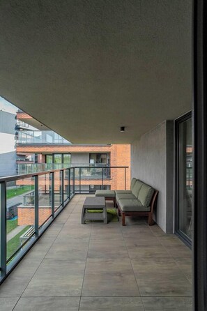 Ein Balkon mit einer Couch, einem kleinen Tisch, einem Glasgeländer und Blick auf ein Backsteingebäude.