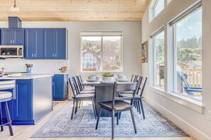 Eine moderne Küche mit blauen Schränken, ein Essbereich mit Tisch für vier Personen und große Fenster mit Blick auf eine Wohngegend.