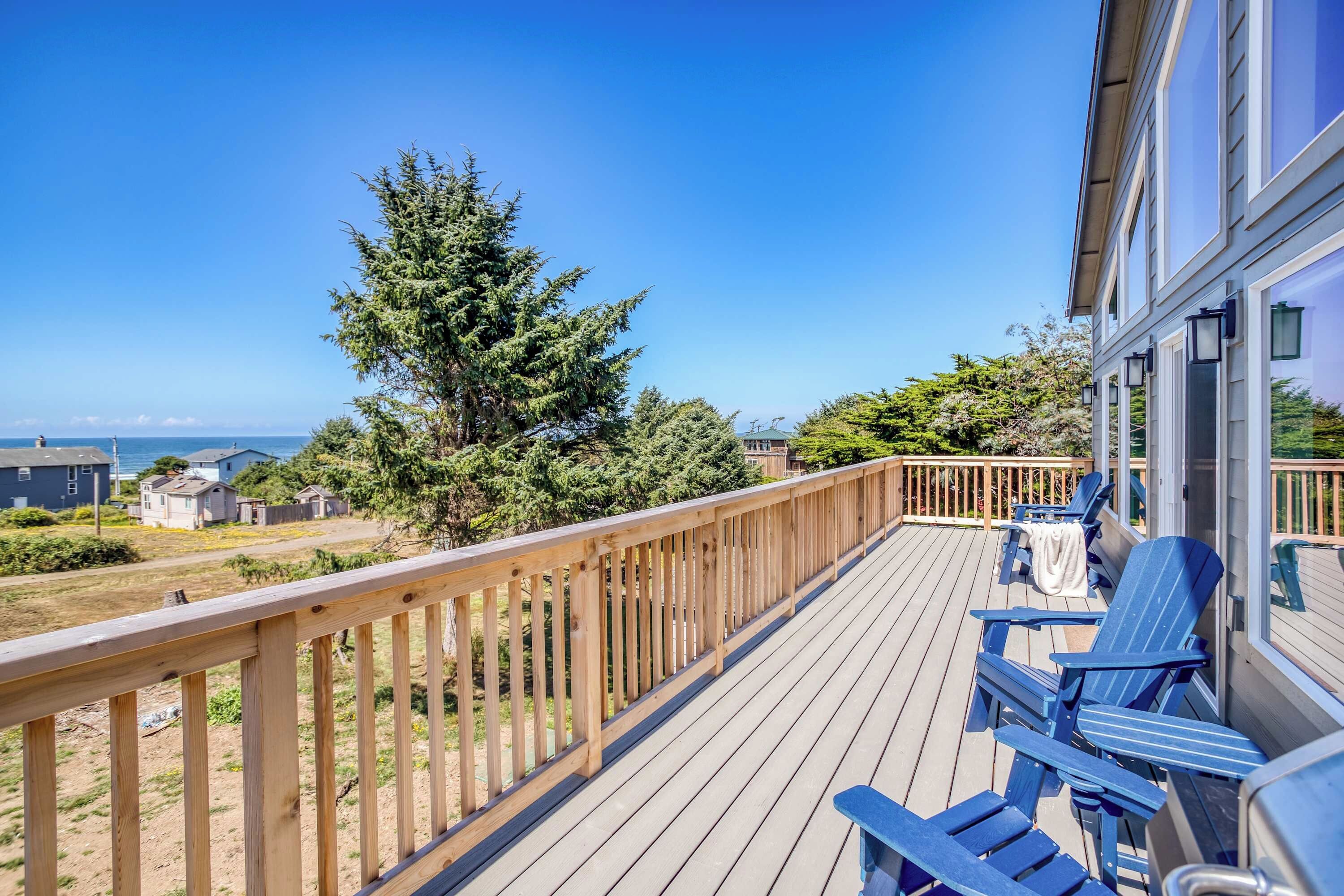 Une terrasse en bois avec des chaises bleues, donnant sur une zone côtière avec des maisons et un grand arbre à feuilles persistantes.