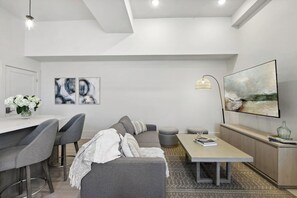A modern living room with a grey sofa, a dining table, and a coffee table. There are two framed abstract paintings on the wall and a large landscape painting above the coffee table. - Coastal Retreat, Downtown Bay St Louis (Bay St. Louis)
