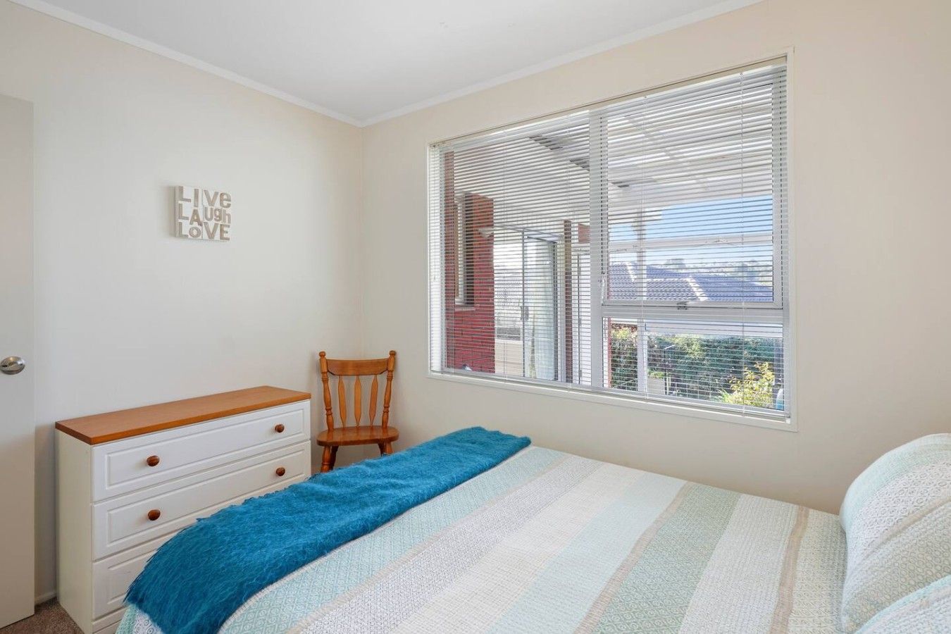 A bedroom with a bed, a wooden dresser, a chair, and a window with blinds.