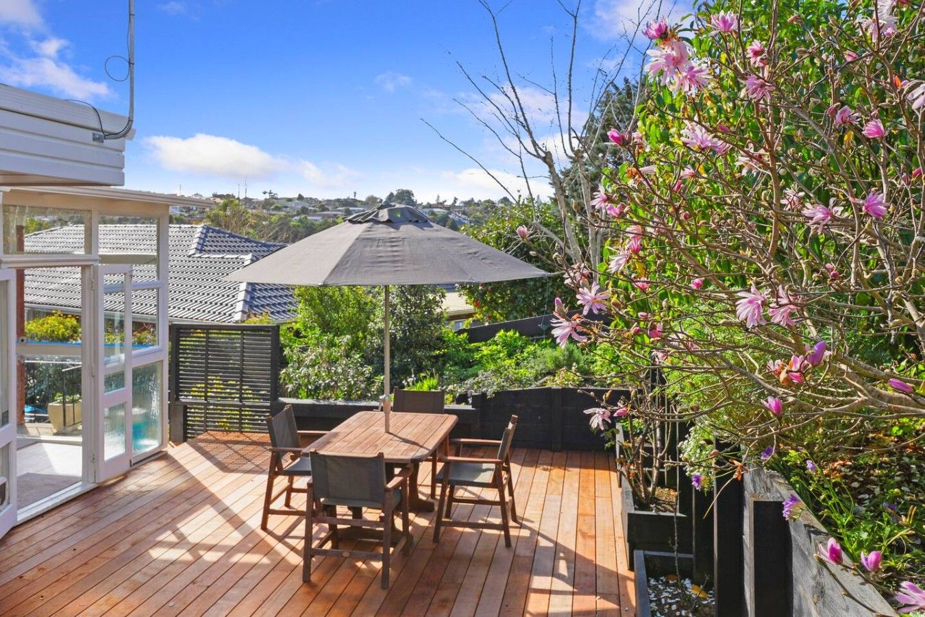 A wooden deck with a table and chairs, surrounded by greenery and a flowering tree.