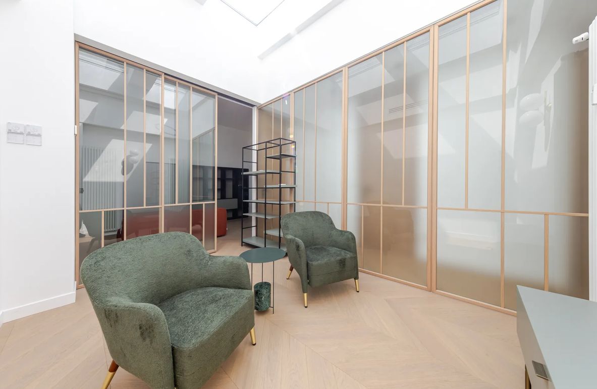 A modern room with a glass partition, two green armchairs, a small round table, and a metal shelving unit.
