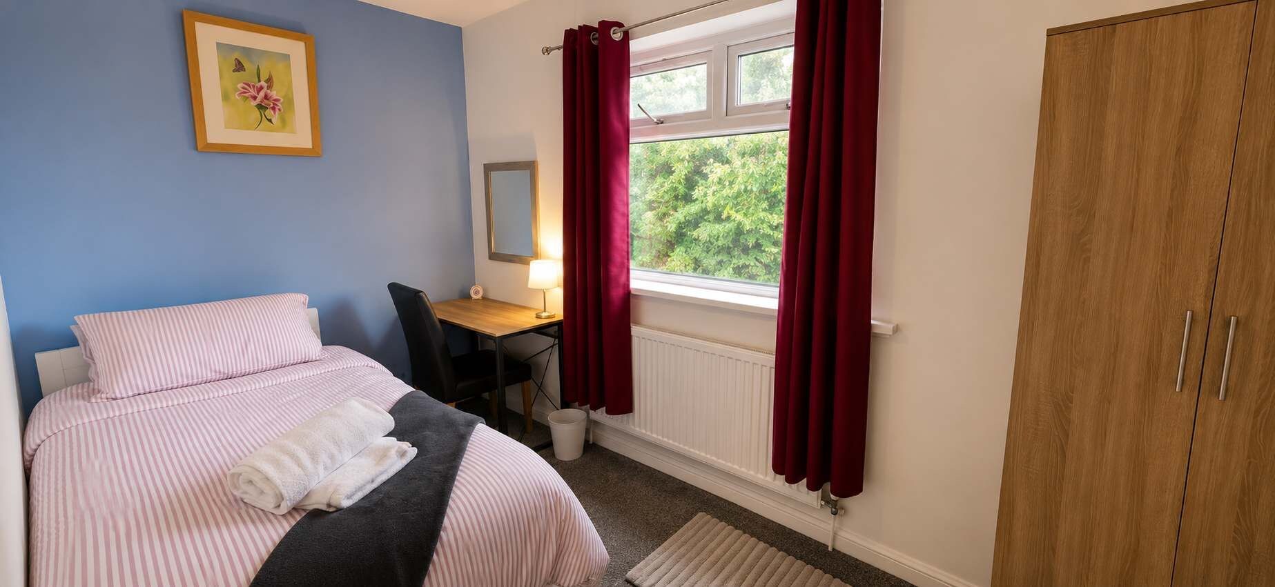 A bedroom with a bed, a desk, a chair, a wardrobe, and a window with curtains.