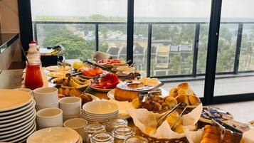 Uma mesa de buffet com diversos alimentos e pratos empilhados, com o cenário de uma varanda e vista para edifícios e vegetação.