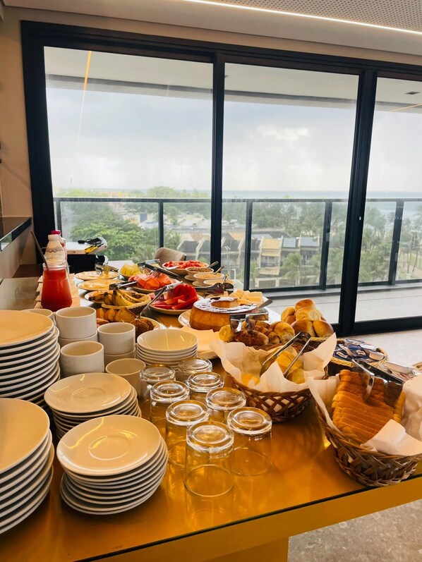 A buffet table with various food items and stacked plates, set against a backdrop of a balcony with a view of buildings and greenery.