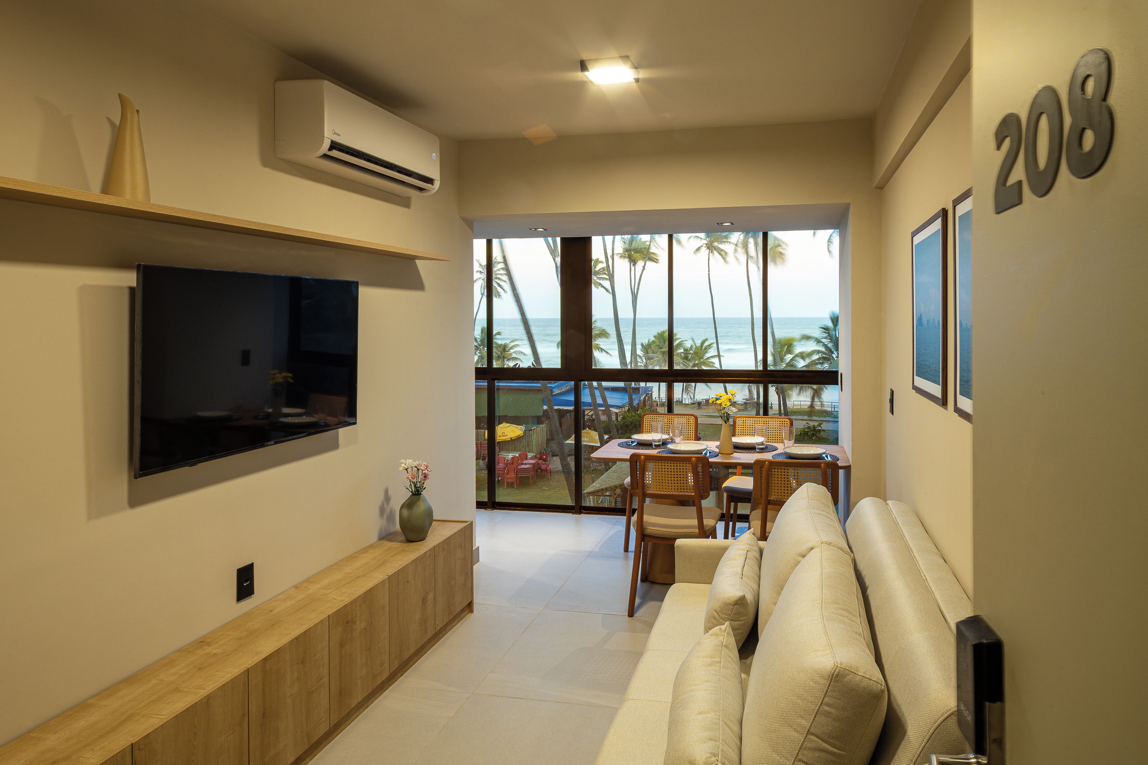A modern living room with a flat-screen TV, a sofa, a dining area with chairs and a table, and a large window with a view of palm trees and the ocean.