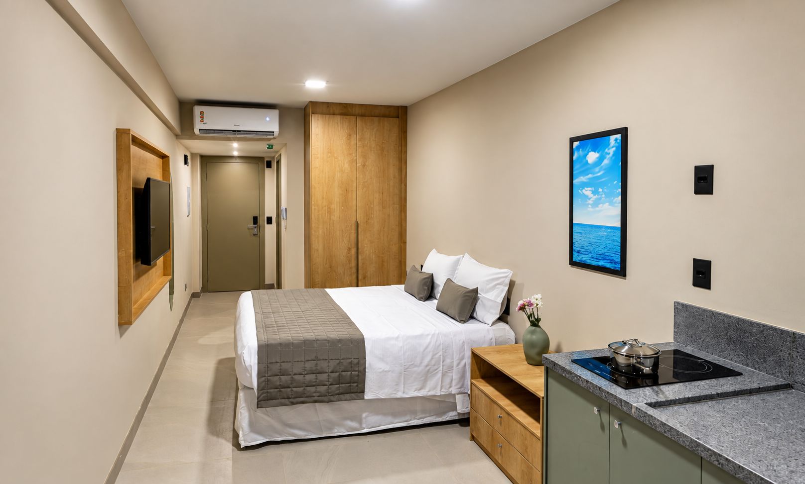 Una habitación de hotel moderna con una cama, un televisor de pantalla plana, un cuadro, una zona de cocina con estufa y un armario de madera.