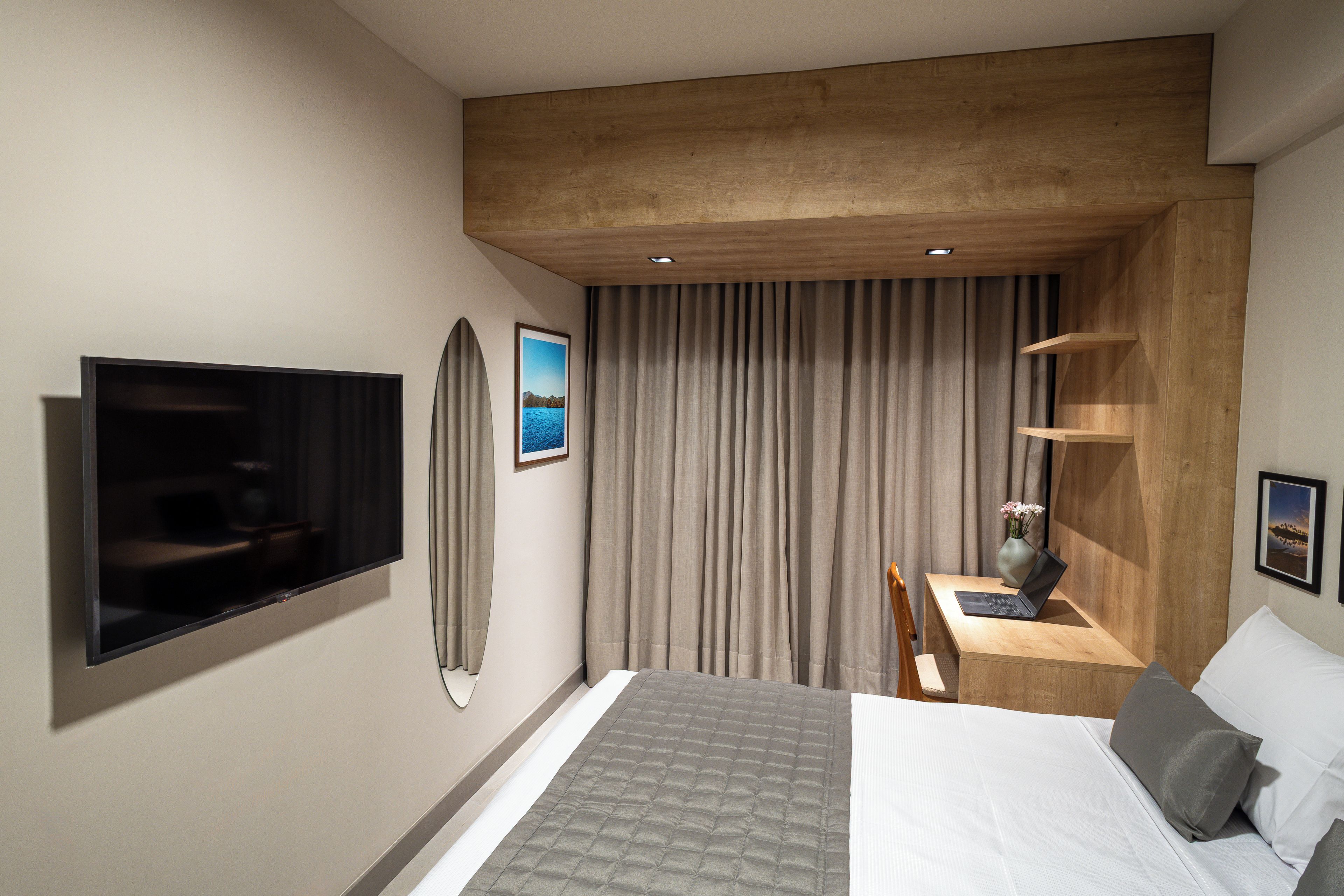 Una habitación de hotel moderna con una televisión de pantalla plana, una cama con colcha gris, un escritorio con una computadora portátil y un ventanal con cortinas.