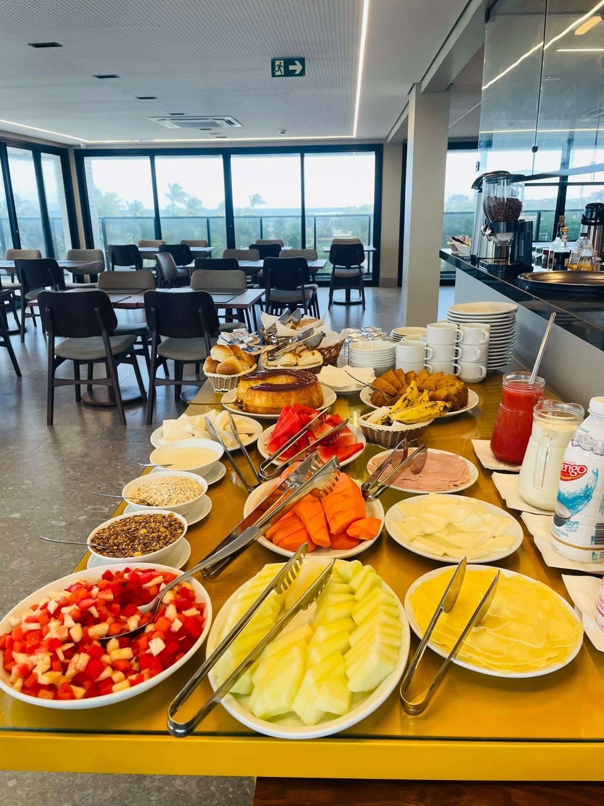 A buffet table with various food items and utensils, a coffee station, and a view of the outdoors through large windows.