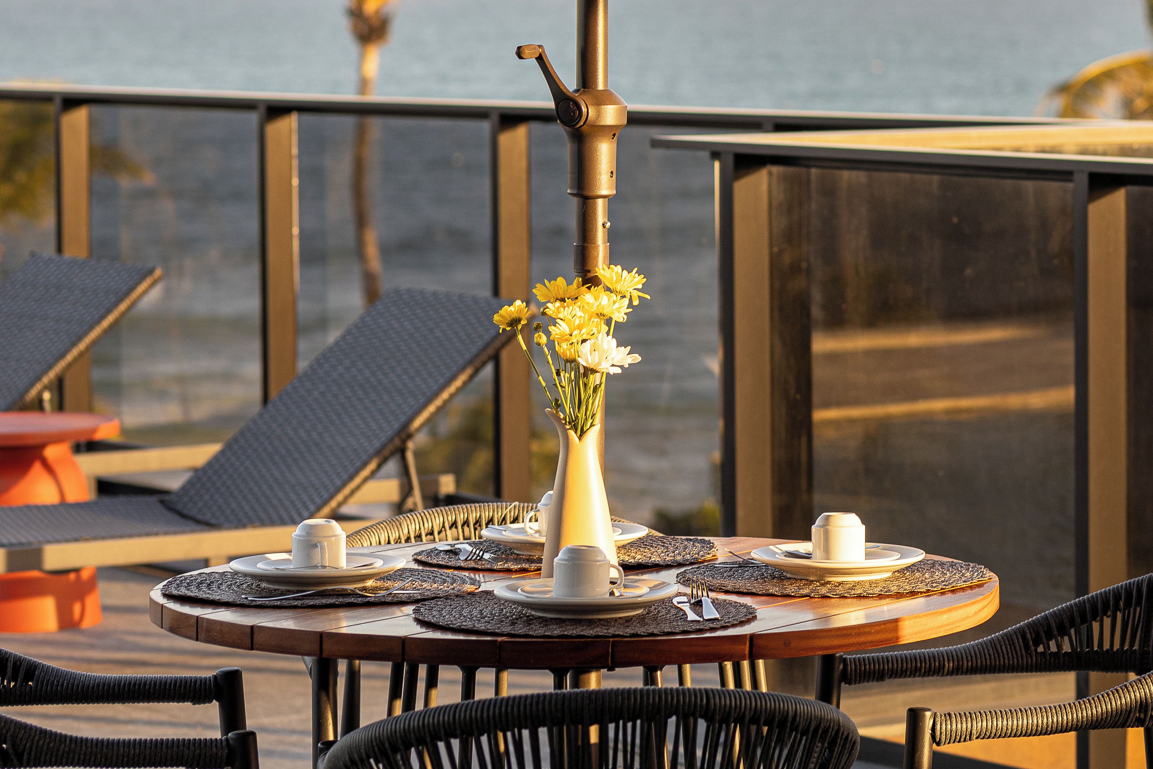 Una mesa redonda con un florero de flores amarillas, tazas blancas y un candelabro, colocada en un balcón con sillas y vista al mar.