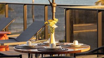Una mesa redonda con un florero de flores amarillas, tazas blancas y un candelabro, colocada en un balcón con sillas y vista al mar.