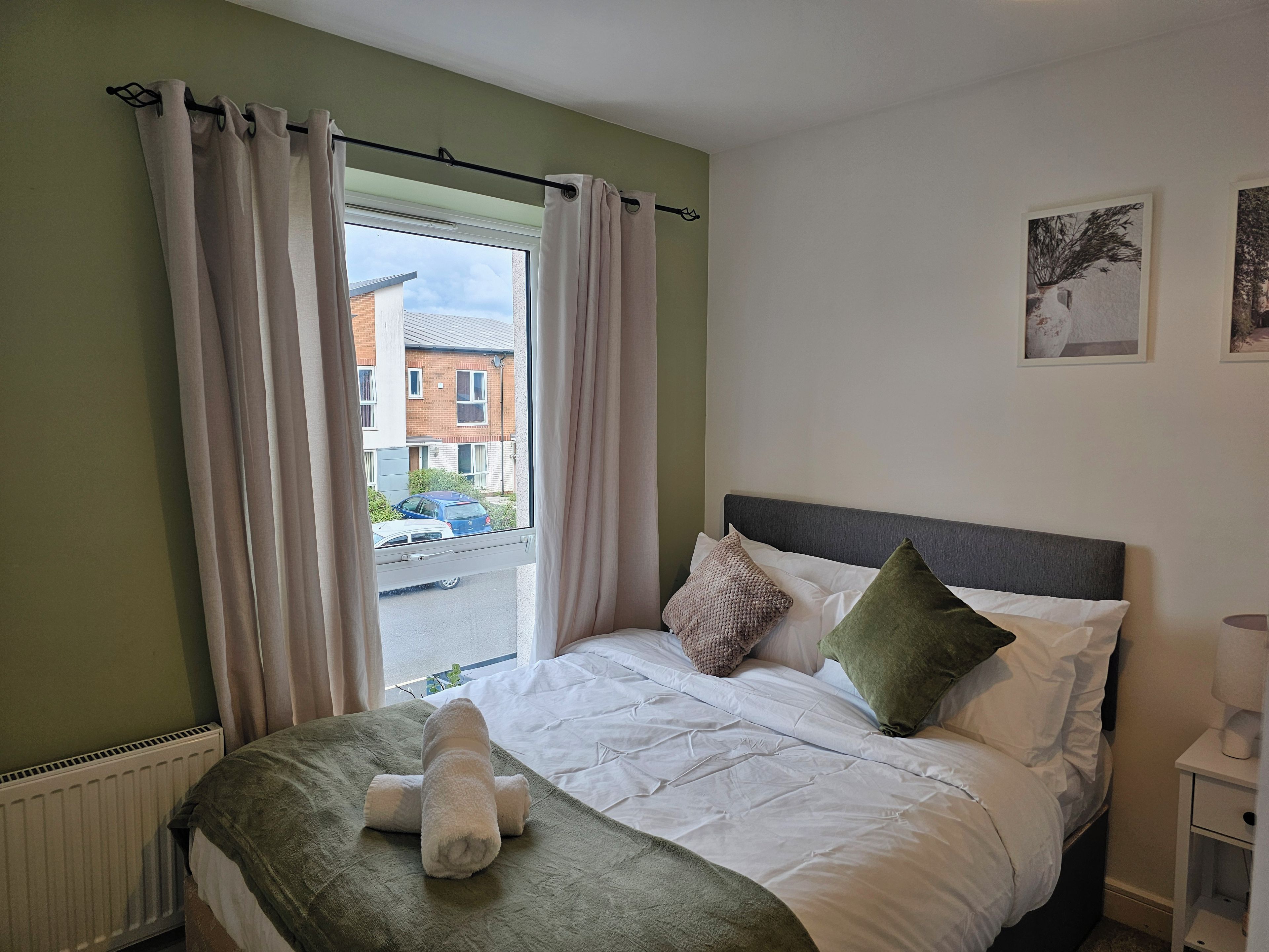 A bedroom with a bed, pillows, a bedside table, a radiator, and a window with curtains.