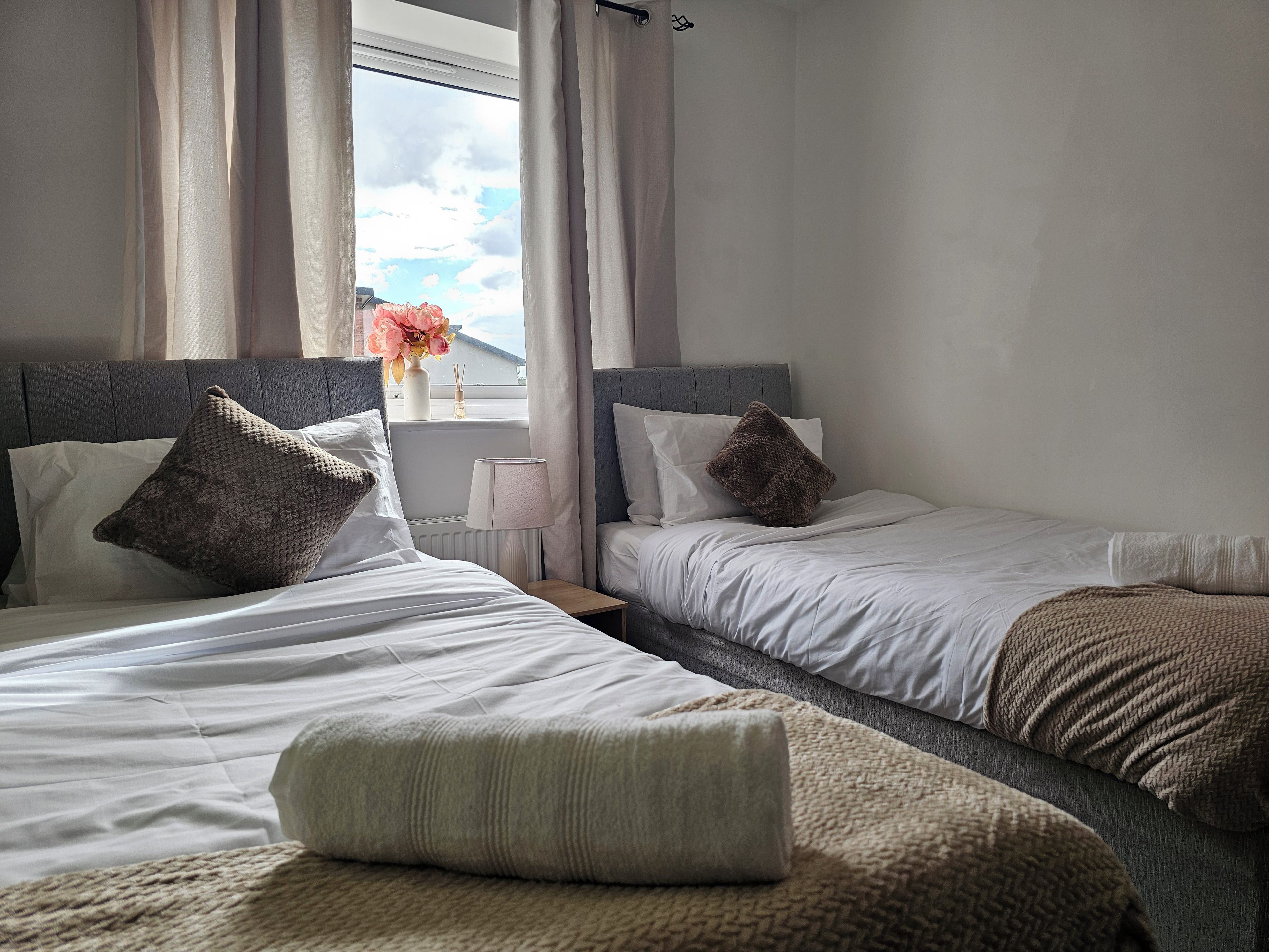 Habitació d'hotel amb dos llits, una finestra amb cortines, una taula de nit amb una làmpada i vista al cel.