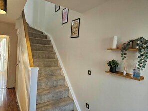 Una escalera con peldaños alfombrados y barandilla de madera, que conduce a un rellano con interruptores de luz y cuadros en la pared.