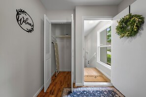 Ein Flur mit einem 'Welcome'-Schild, einer Tür zu einem Zimmer, einem Fenster mit Blick und einer Fußmatte mit einem Namen.