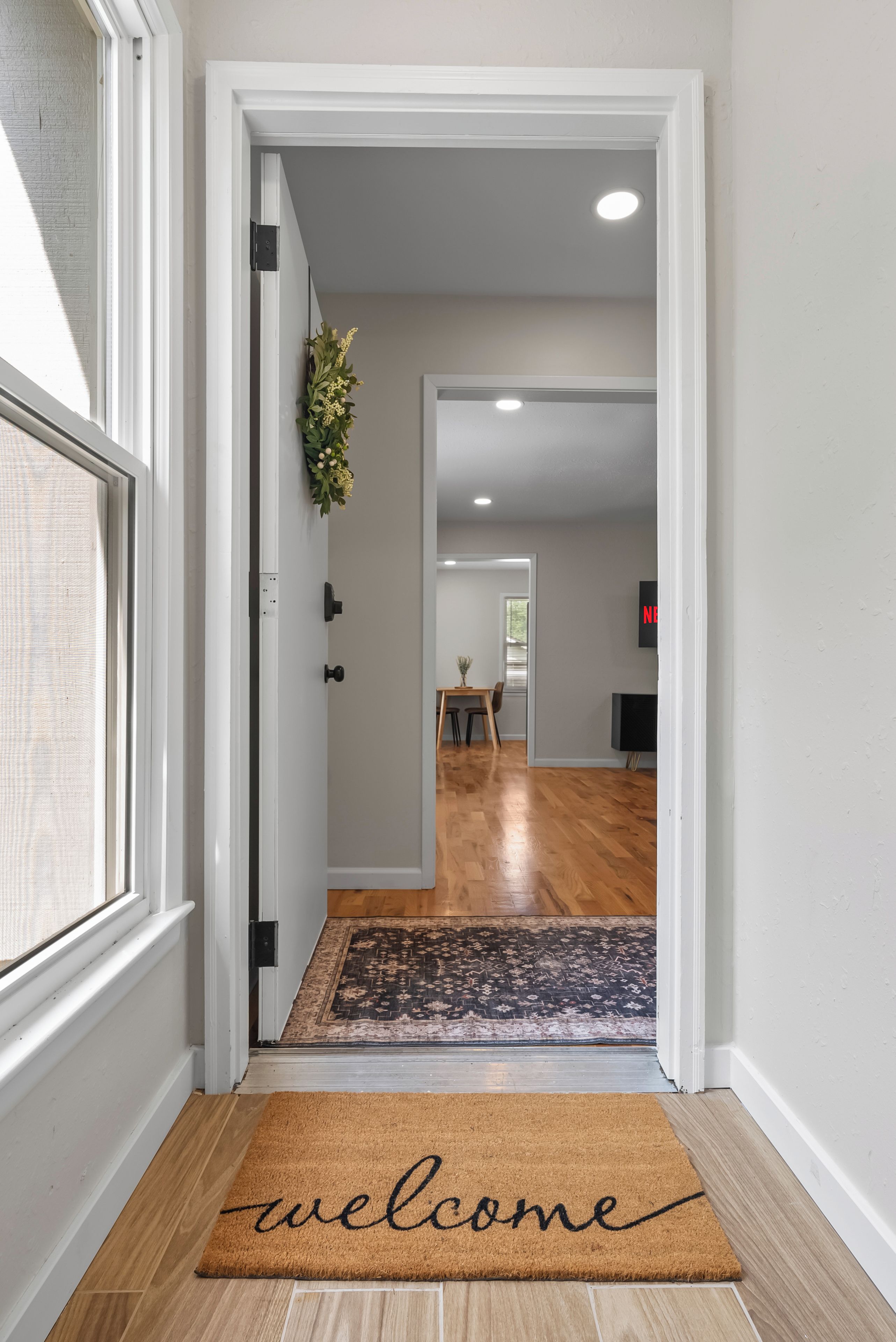 A hallway with a welcome mat, a door, a plant, and a view into a room with a table and chairs.