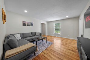 Ein modernes Wohnzimmer mit einem grauen Ecksofa, einem Couchtisch und einem Fernseher auf Holzdielen.