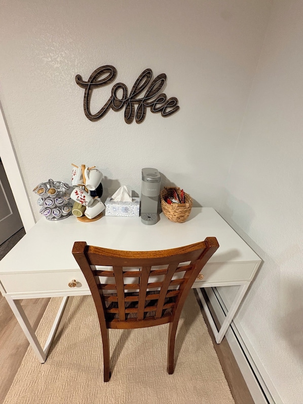 A white desk with a wooden chair, a wall-mounted sign that reads 'Coffee', and various items including a mug, a tissue box, and a basket.