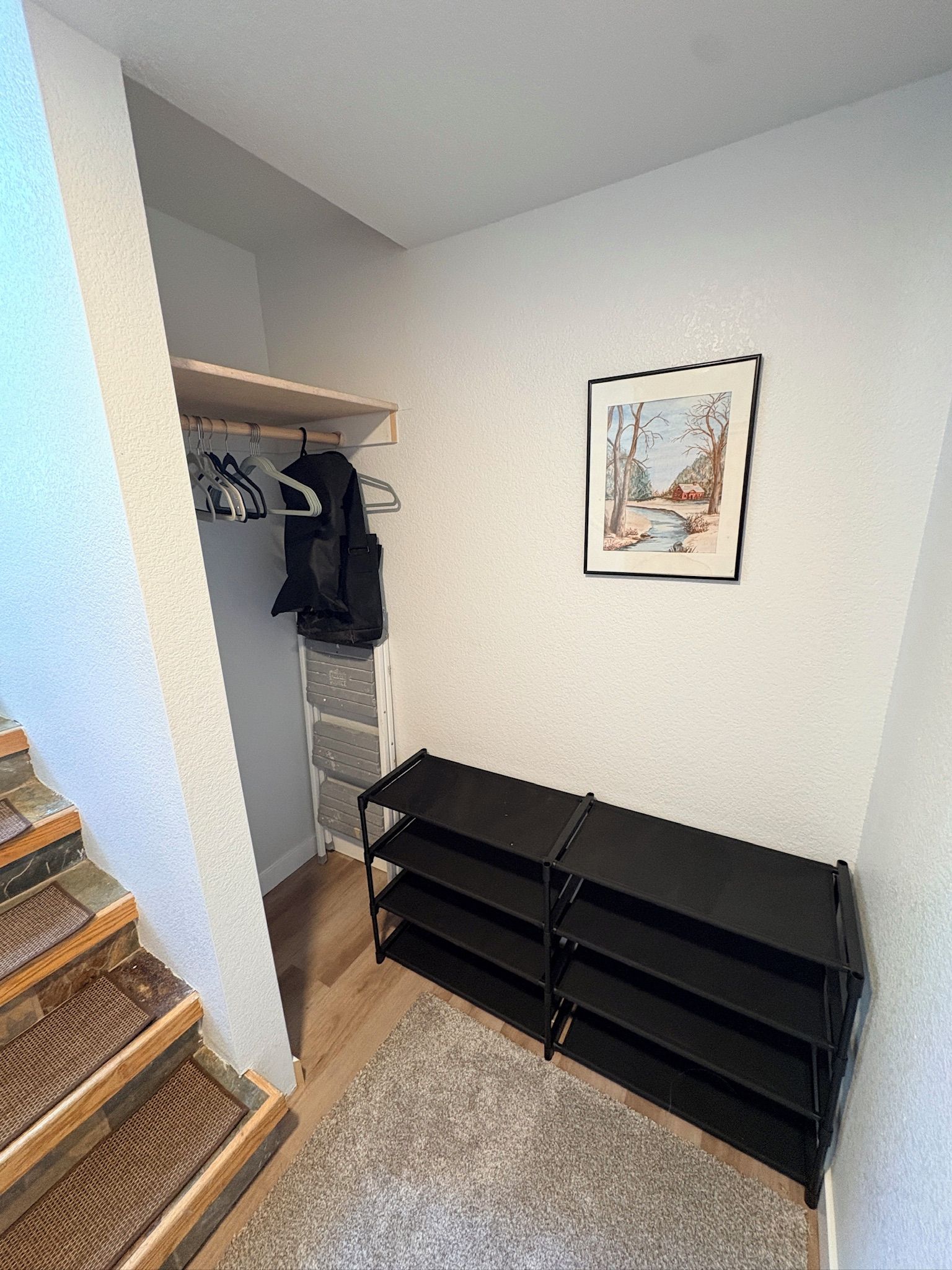 A room with a wooden staircase, a black metal shelf unit, a coat rack with hanging clothes, and a framed picture on the wall.
