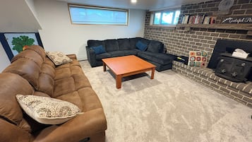 Een woonkamer met een bruine bank, een houten salontafel, een donkergrijze hoekbank en een schouw van baksteen.
