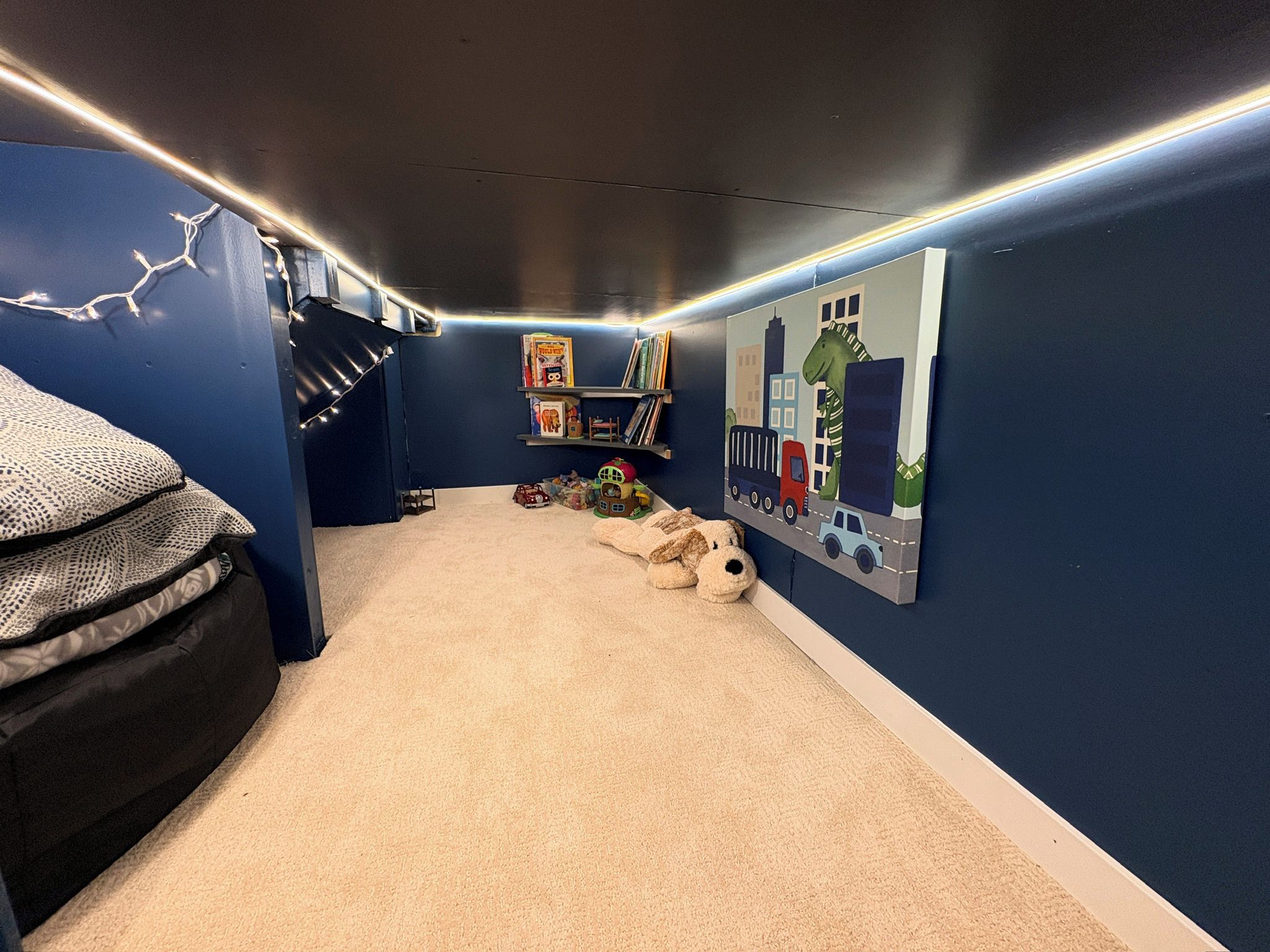 A room with a carpeted floor, a bed with a patterned cover, a bookshelf with books and toys, and a wall with a cityscape mural.