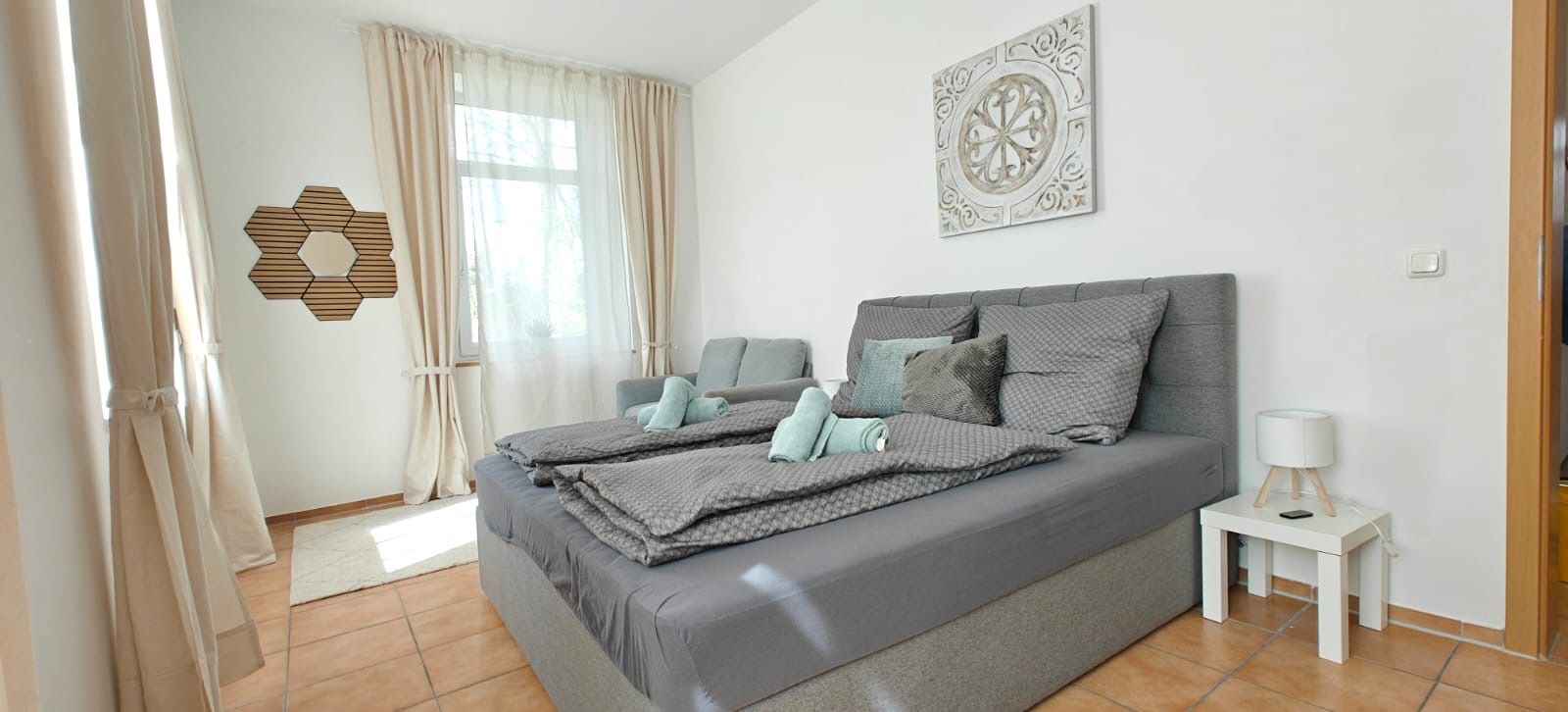 A bedroom with a bed, a nightstand, a lamp, a decorative wall piece, and a window with curtains.