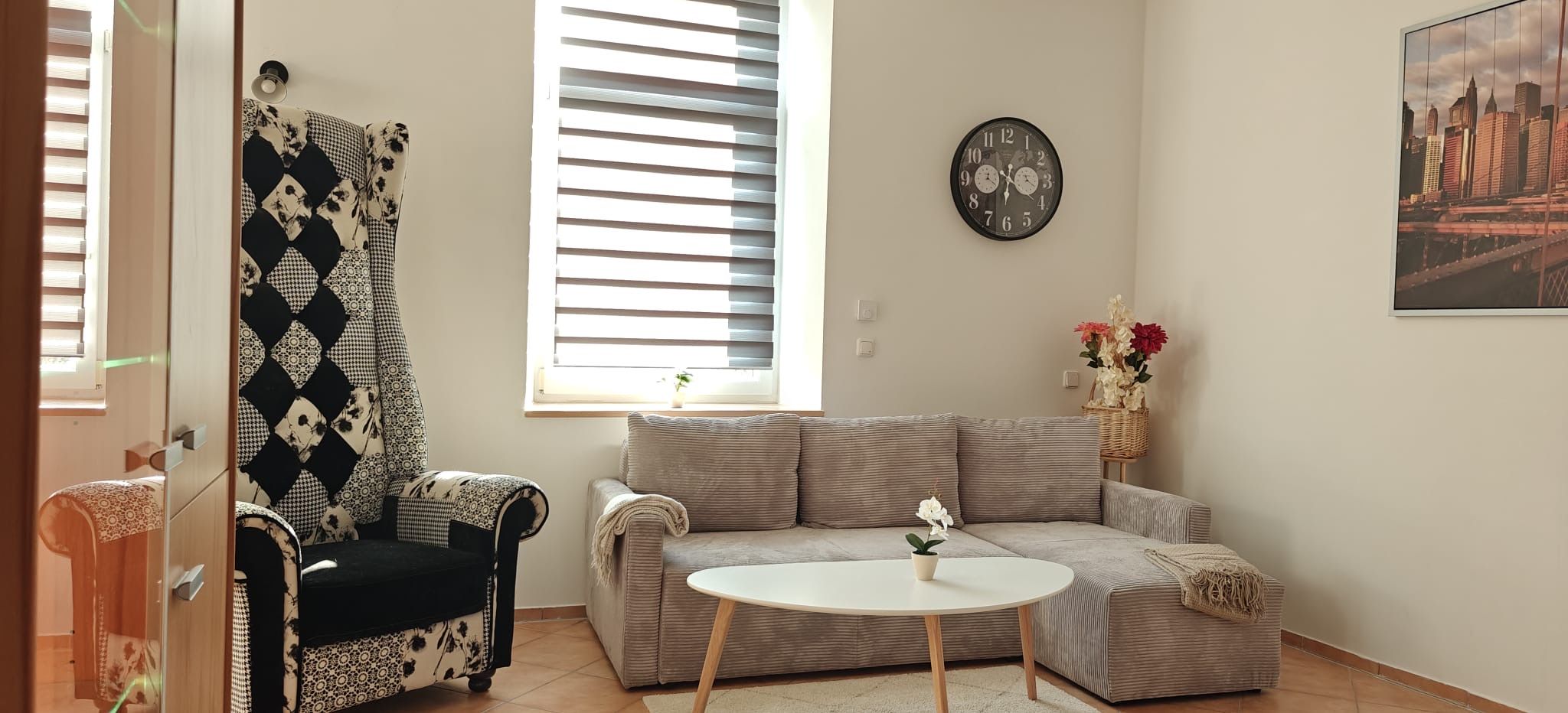 Ein Wohnzimmer mit einer Couch, einem Sessel, einem runden Tisch, einer Uhr und einem Fenster mit Jalousien.