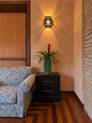 Quarto com piso de madeira, sofá estampado, mesa de cabeceira pequena preta com gavetas e luminária fixada na parede.