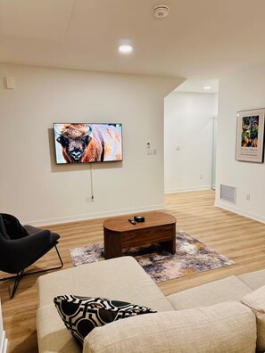 A modern living room with a flat-screen TV mounted on the wall, a wooden coffee table, a black armchair, a beige sofa, and a patterned rug. - Modern & Cozy Townhouse - 2 Bed + 3 Bath w/Parking (Brampton)
