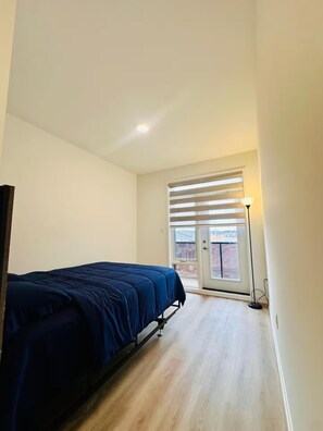 Ein Schlafzimmer mit einem Bett, einem Fenster mit Jalousien, einer Stehlampe und Blick auf Gebäude.
