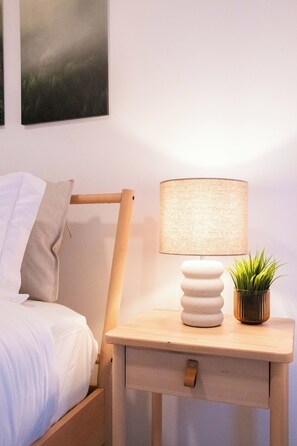 A neatly made bed with white linens, a wooden nightstand with a lamp and a small potted plant, and a wall-mounted painting above the bed. - NOK Tropical 1BR Escape in Vistacana (Punta Cana)