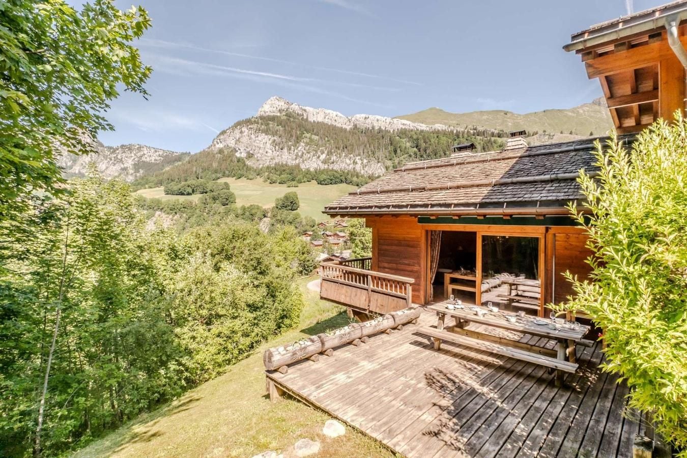 A wooden cabin with a deck and outdoor seating, surrounded by lush greenery and mountains in the background.