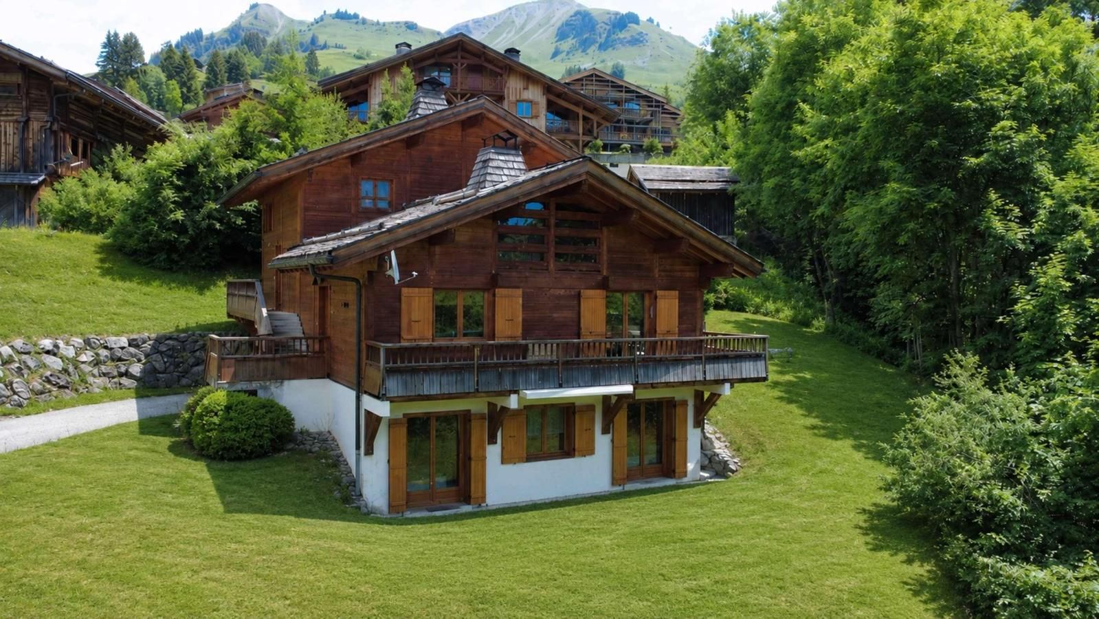 Un chalet de madera de dos pisos con un balcón, rodeado de exuberante vegetación y montañas al fondo.