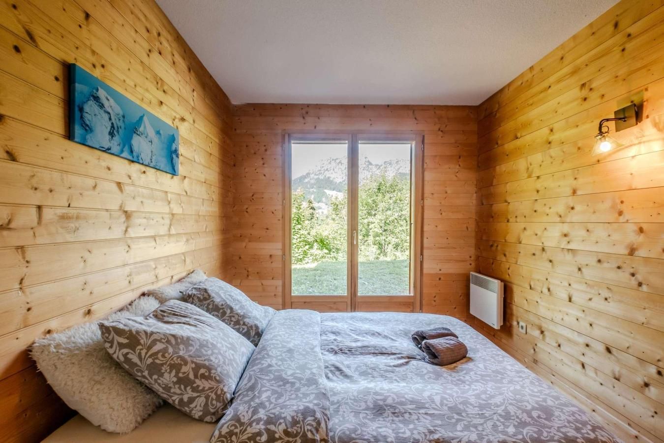 A wooden-paneled room with a bed, pillows, a painting, and a window with a view of greenery.