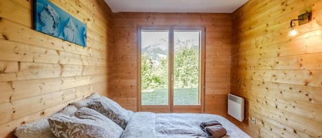 Ett rum med träpanel på väggarna, en säng, kuddar, en tavla och ett fönster med utsikt över grönskan.